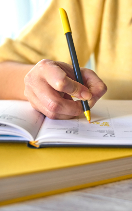 Young person in a yellow shirt writing in a notebook with a yellow pen